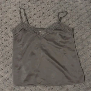 Grå linnetopp med spets från Kappahl - Säljer en grå linnetopp från Kappahl med tunna axelband och vacker spetsdetalj längs v-ringningen. Toppen har en mjuk satinliknande yta och är perfekt för att skapa en stilren look.