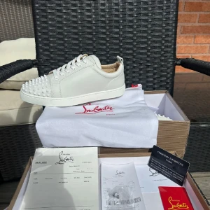 Vita sneakers med nitar från Christian Louboutin - Intresse koll på dessa vita sneakers från Christian Louboutin med ikoniska röda sulor och silverfärgade nitar framtill. Skorna har snörning och är tillverkade i skinn. Perfekta för dig som vill sticka ut med en lyxig och edgy stil. Endast seriösa köpare. Kan gå ner i pris vid smidig affär.