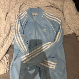 Ljusblå Adidas track jacket - Säljer en ljusblå Adidas track jacket med vita ränder längs ärmarna och dragkedja framtill. Klassisk retrostil med ribbade muddar och Adidas-logga på bröstet. Perfekt för en sportig och avslappnad look. Addidas sst, somrig färg 