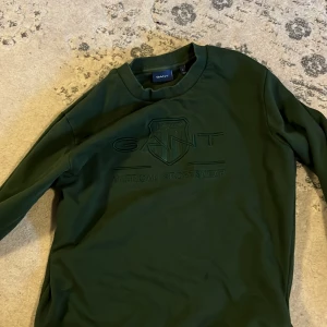 Mörkgrön sweatshirt från GANT - Säljer en mörkgrön sweatshirt från GANT med broderad logga och text framtill. Tröjan har rund halsringning och långa ärmar. Perfekt för en avslappnad och stilren look.