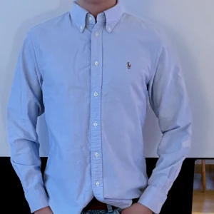 Ljusblå skjorta från Ralph Lauren - Klassisk ljusblå skjorta från Ralph Lauren med button-down krage och broderad logga på bröstet. Skjortan har långa ärmar och vita knappar framtill. Perfekt till jeans eller chinos för en stilren look.