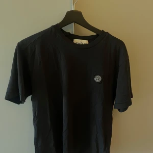 Svart t-shirt från Stone Island - Säljer en svart t-shirt från Stone Island med klassisk logotyp-patch på bröstet. T-shirten har rund hals och är tillverkad i mjuk bomull. Perfekt för en stilren och enkel look.