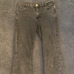 Svarta jeans med slitning - Säljer ett par gråa lågmidjade bootcut jeans med lätt slitning på ena benet. Jeansen har klassisk femficksdesign, raka ben med bootcut. Stängs med knapp och dragkedja. Perfekta till vardags och enkla att matcha med olika toppar.