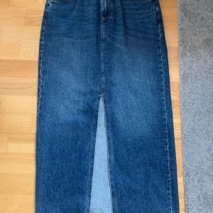Jeanskjol i klassisk blå denim - Säljer en stilren blå jeanskjol med rak passform och slits framtill. Kjolen har klassiska bakfickor och är i en längre modell som passar perfekt till sneakers eller boots. Enkel och tidlös design som är lätt att matcha.