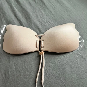 Beige snörnings-bh axelbandslös SJÄLVHÄFTANDE BH bra  - Säljer en beige bh  med snörning framtill och transparenta axelband. Kuporna har en slät utsida och insidan är försedd med små pluppar för bättre grepp. Perfekt för dig som vill ha en diskret look under sommaren.