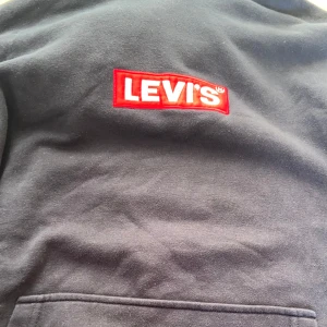Svart hoodie från Levi's - Säljer en svart hoodie från Levi's med den klassiska röda loggan broderad på bröstet. Tröjan har en stor ficka framtill och är perfekt för en avslappnad stil. Passar dig som gillar streetwear och vill ha något bekvämt och snyggt.