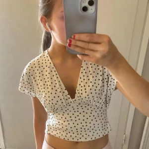 Vit prickig croppad blus från Zara - Säljer en vit croppad blus från Zara med svarta prickar. Blusen har djup v-ringning, korta ärmar och smockad midja som ger en snygg passform. Perfekt till sommaren och lätt att matcha med både jeans och kjol.