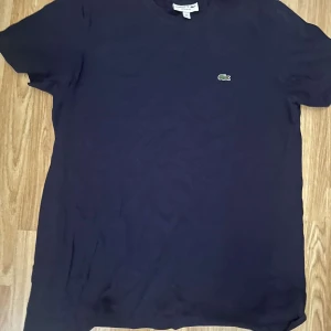 Mörkblå t-shirt från Lacoste - Säljer en klassisk mörkblå t-shirt från Lacoste med den ikoniska broderade krokodilloggan på bröstet. T-shirten har rund hals och korta ärmar, perfekt för en clean och stilren look.