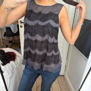 Grå ärmlös blus med spetsdetaljer - Supersnygg grå ärmlös blus med transparent mesh upptill och vågiga spetsdetaljer framtill. Blusen har en luftig passform och knäppning i nacken. Perfekt att styla med jeans eller kjol.