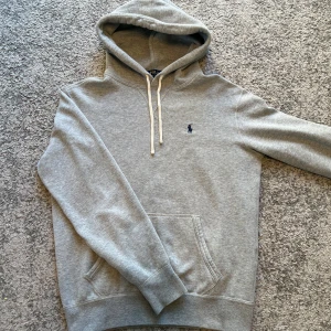 Ralph Lauren hoodie  - Säljer en klassisk grå hoodie från Polo Ralph Lauren med broderad marinblå logga på bröstet. Tröjan har huva med dragsko och en stor magficka. Perfekt för en avslappnad och stilren look.