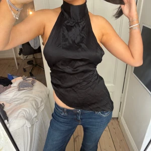 Svart halterneck topp med rosett - Säljer en svart halterneck topp med hög krage och knytning i nacken som ger en snygg rosett baktill. Toppen är ärmlös och har en slät, glansig yta som känns lyxig. Perfekt till jeans eller kjol för en trendig look.