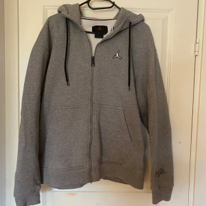 Grå hoodie från Jordan med dragkedja - Säljer en grå hoodie från Jordan med dragkedja och huva. Tröjan har svarta snören, broderad logga på bröstet och en signaturdetalj på ärmen. Perfekt för en sportig och avslappnad stil.