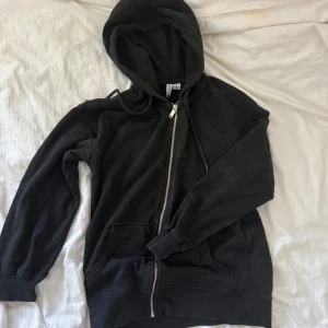 Svart hoodie med dragkedja  - Svart oversized hoodie med dragkedja framtill och huva med snörning. Tröjan har långa ärmar, ribbade muddar och två stora fickor framtill. Perfekt för chill dagar eller när du vill ha en enkel och stilren look.