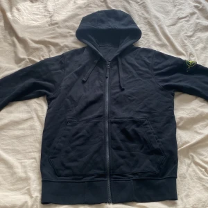 Stone Island huvtröja - Svart hoodie från Stone Island med klassisk logga på ärmen. Tröjan har dragkedja framtill, huva med snörning och två stora fickor. Tillverkad i mjuk bomull, perfekt för en chill och stilren look.