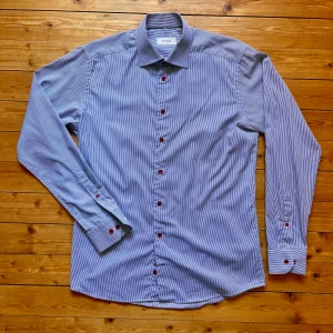 Randig blå skjorta från Eton slim fit - Snygg blå- och vitrandig skjorta från Eton i slim fit-modell. Skjortan har röda knappar som ger en cool kontrast och klassisk krage. Tillverkad i bomull och har långa ärmar med dubbla knappar vid manschetten. Perfekt för dig som gillar stilrena detaljer.
