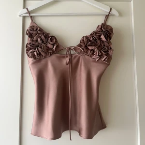 Rosé satin linnetopp med volang - Slutsåld supersnygg rosé  linnetopp i satin med tunna axelband och volangdetaljer över bysten. Toppen har en söt knytning framtill och en lätt glansig finish som ger en lyxig känsla. Perfekt för dig som vill sticka ut med något extra. Linnet är litet i modellen så skulle även passat en S. Använd en gång. 