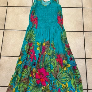 Turkos långklänning med tropiskt mönster - Färgstark långklänning i turkos med tropiskt blommönster och papegojor i rosa, gul, blå och grön. Smockad överdel och ärmlös design, perfekt för varma dagar. Lätt och luftig känsla, tillverkad i bomull. Snyggt flowy fall och riktigt somrig vibe!