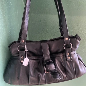 Svart axelväska i skinnlook - Snygg svart axelväska med två långa remmar och silverfärgade metalldetaljer. Väskan har ett spänne framtill och dragkedja på baksidan. Tillverkad i skinnimitation med mjuk, lätt rynkad form. Perfekt för dig som vill ha en stilren och praktisk väska.