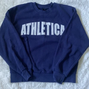 Blå ATHLETICA sweatshirt från Gina Tricot - Mörkblå sweatshirt från Gina Tricot med stor vit ATHLETICA-text framtill. Klassisk rund halsringning, ribbade muddar och lång ärm. Skön och avslappnad passform i mjuk bomull, perfekt för en sportig look.