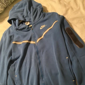 Blå Nike Tech Tröja - Nike Tech Tröja i storlek 158-170. Jackan har hel dragkedja, huva och vita detaljer över bröstet. Ficka med dragkedja på ena ärmen,  två sidofickor och två innerfickor. Storlek 158-170 i fint skick
