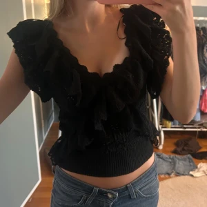 Svart volangblus med v-ringning - Supersöt svart blus med djup v-ringning och stora volanger längs axlar och halsringning. Toppen är croppad och har ribbad midja som sitter snyggt. Perfekt för dig som gillar romantiska detaljer och vill sticka ut lite extra.