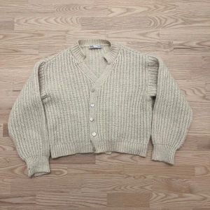 Beige stickad kofta från Zara - Supermysig beige stickad kofta från Zara med v-ringning och stora silvriga knappar. Koftan har en chunky struktur, ribbade muddar och lång ärm. Perfekt att slänga på sig när det är kyligt ute.