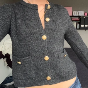 Mörkgrå stickad kofta med guldknappar - Snygg mörkgrå stickad kofta med guldiga knappar framtill och på fickorna. Croppad modell med lång ärm och två fickor framtill. Perfekt för dig som gillar en klassisk men trendig look.