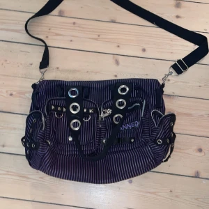 Randig axelväska med nitar och dragkedjor - Cool axelväska från Banned i svart och lila med smala ränder. Väskan har flera dragkedjor, stora silverfärgade öljetter och detaljer i tyg. Justerbar axelrem och extra handtag med nitar ger en edgy vibe. Perfekt för dig som gillar alternativ stil och vill sticka ut. En dragkedja är sönder men går att fixa hos skräddare om man vill