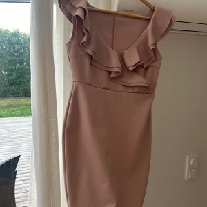 Ljusrosa kort klänning med volang storlek M från Nelly - Supersöt ljusrosa kort klänning med volangdetaljer längs axel och byst. Klänningen har en figurnära passform och är ärmlös med breda axelband. Perfekt för dig som gillar en feminin och stilren look.