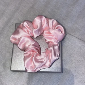 Rosa scrunchie i glansig satin - En superfin scrunchie i ljusrosa satin som ger en lyxig känsla till din frisyr. Den är fluffig och glansig, perfekt för att piffa upp håret med en söt detalj. Passar både till en enkel tofs eller runt en knut för extra volym. 