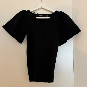 Svart topp med puffärm från Vero Moda - Stilren svart topp från Vero Moda med v-ringning och stora puffärmar som ger en trendig siluett. Toppen är figurnära och har korta ärmar med extra volym. Perfekt för dig som vill sticka ut med en enkel men cool look.