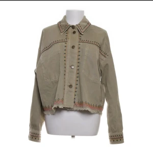 Beige broderad jeansjacka med frans - Snygg beige jeansjacka med broderade detaljer i flera färger längs krage, ärmslut och nederkant. Jackan har knappar framtill, två stora fickor och rå fransad nederkant för en chill vibe. Perfekt för dig som gillar unika plagg med boho-touch.