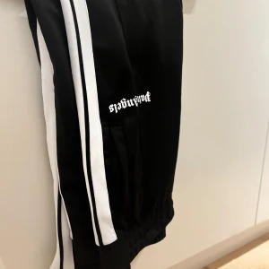 Palm Angels svarta mjukisbyxor med revärer - Svarta mjukisbyxor från Palm Angels med vita revärer längs sidorna och logga tryckt på låret. Byxorna har dragkedjor vid bensluten och är gjorda i ett mjukt syntetmaterial. Perfekta för en sportig och trendig look.