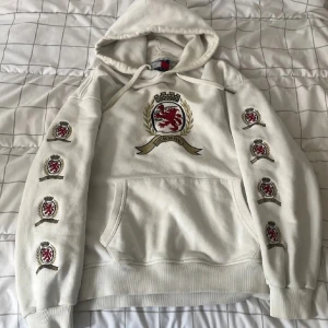 Vit hoodie Tommy Jeans med brodyr - Vit hoodie från Tommy Jeans med broderade emblem i rött, guld och svart på bröstet och längs båda ärmarna. Klassisk huva med snörning och stor magficka framtill. Skön passform och snygga detaljer som ger en preppy vibe.