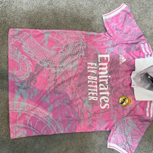 Real Madrid rosa fotbollströja Adidas - Snygg Real Madrid fotbollströja från Adidas i rosa med grått drakmönster. Tröjan har vit krage, korta ärmar och tryck med Emirates Fly Better på bröstet. Materialet är lätt och andas, perfekt för match eller träning. Real Madrid-logga och Adidas-märke på framsidan.