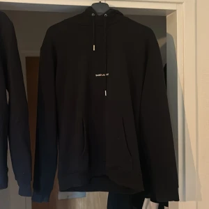 Svart Saint Laurent hoodie med logga - Svart hoodie från Saint Laurent med vit och röd broderad logga på bröstet. Klassisk huva med snörning och känguruficka framtill. Mjuk bomullskänsla och långärmad modell som ger en clean och stilren vibe.