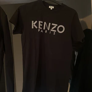 Svart Kenzo t-shirt med logga - Svart t-shirt från Kenzo med stor vit logga 'KENZO PARIS' på bröstet. Klassisk rund halsringning och korta ärmar. Tillverkad i mjuk bomull som känns skön mot huden. Perfekt för dig som gillar stilrena statement-plagg.