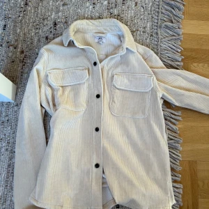 Beige manchester overshirt - Snygg beige overshirt i manchester med svarta knappar och två stora bröstfickor. Skjortan har lång ärm och klassisk krage, perfekt att slänga över en t-shirt för en chill look.