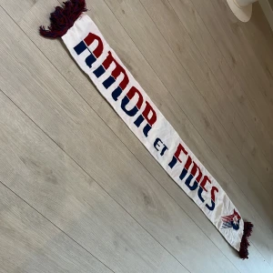 ÖIS halsduk i rött, blått och vitt - Supporterhalsduk från ÖIS med texten 'Amor et Fides' i rött och blått på vit botten. Halsduken har broderad ÖIS-logga och matchande röda och blå fransar i ändarna. Perfekt accessoar för att visa ditt stöd på läktaren.