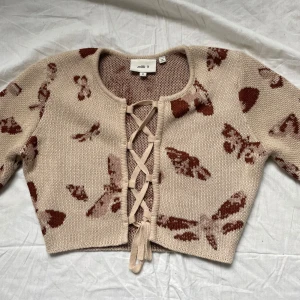 Beige kofta med fjärilsmönster och snörning, Milk It - Superfin croppad beige kofta från Milk It Vintage med brunt fjärilsmönster och snygg snörning framtill. Koftan är stickad, har långa ärmar och rund halsringning. Lite tjockare material. Perfekt för dig som gillar unika detaljer och trendiga plagg. Aldrig använd pga för liten. 