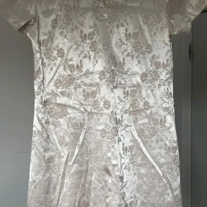 Champagnefärgad blommig kortklänning - Snygg kortklänning i champagnefärgat satin med diskret blommigt mönster. Klänningen har korta ärmar och en elegant, glansig finish som ger en lyxig känsla. Perfekt för dig som vill sticka ut med en stilren och trendig look.