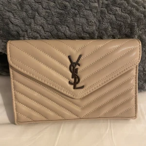 Beige YSL quiltad axelväska i skinn - Säljer en beige quiltad axelväska från YSL med klassiskt YSL-emblem i metall framtill. Väskan har kedjerem och är tillverkad i skinn med snygga diagonala sömmar. Perfekt storlek för det viktigaste och ger en lyxig touch till din outfit.