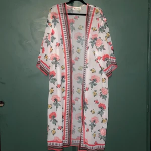 Blommig kaftan H&M Coachella - Superfin kaftan från H&M Coachella-kollektionen med blommigt mönster i rosa, gul och grönt på vit botten. Lätt och luftig med vida ärmar och snygga bårder i rött och svart längs kanterna. Perfekt för sommarens festivalvibbar!