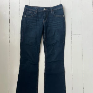 Mörkblå lågmidjade bootcut jeans med broderade fickor - Snygga mörkblå jeans med bootcut passform och klassisk femficksdesign. Baksidan har coola broderade detaljer på fickorna. Jeansen har låg midja och är tillverkade i ett mjukt denimtyg som sitter skönt hela dagen. Är i stl 40 men sitter mer som en 38-36