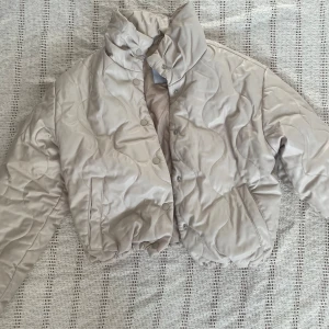 Beige quiltad pufferjacka från Zara - Säljer en beige, quiltad pufferjacka från Zara i storlek XS. Jackan har hög krage, tryckknappar framtill och vågformade sömmar som ger en unik look. Perfekt för dig som gillar en stilren och trendig vibe under kyligare dagar.