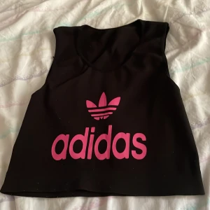 Svart crop top från Adidas - Svart croppad topp  från Adidas med ikoniskt rosa tryck och logga framtill. Toppen är ärmlös och har en sportig vibe, perfekt för träning eller streetwear. Materialet känns stretchigt och bekvämt mot huden.