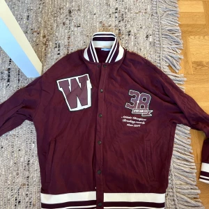 Vinröd bomberjacka med tryck - Snygg vinröd bomberjacka med vita detaljer och tryck framtill. Jackan har stora bokstäver och siffror på bröstet, randiga muddar och krage samt knappar framtill.