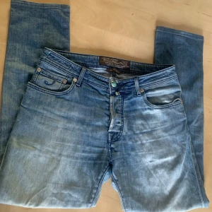 Blå jeans från Jacob Cohën, stl 31 - Snygga blå jeans från Jacob Cohën med klassisk femficksdesign och coolt blått märke bak i midjan. Jeansen har en ljus tvätt och raka ben, tillverkade i mjukt bomullsjeans. Perfekta för dig som gillar stilrena och bekväma jeans.