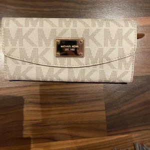 Beige plånbok från Michael Kors - Snygg beige plånbok från Michael Kors med MK-logga i mönstret och guldfärgad metallbricka framtill. Plånboken har flera fack för kort och sedlar, samt dragkedjefack inuti. Utsidan är i tåligt syntetmaterial och insidan är ljusbrun.