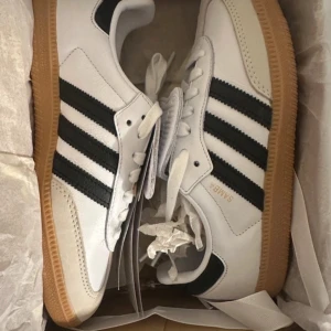 Adidas Samba OG vita och svarta sneakers - Adidas Samba OG sneakers i vitt skinn med svarta klassiska ränder och gummisula. Tungan är extra lång med präglad Adidas-logga. Snygg retrostil med snörning och låg profil, perfekt för dig som gillar streetwear och tidlös design.